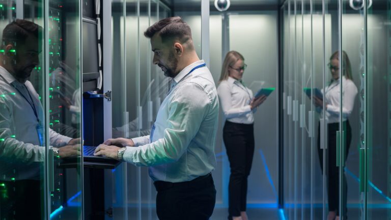 Man typing on laptop in server room & women looking at ipad in backgroud