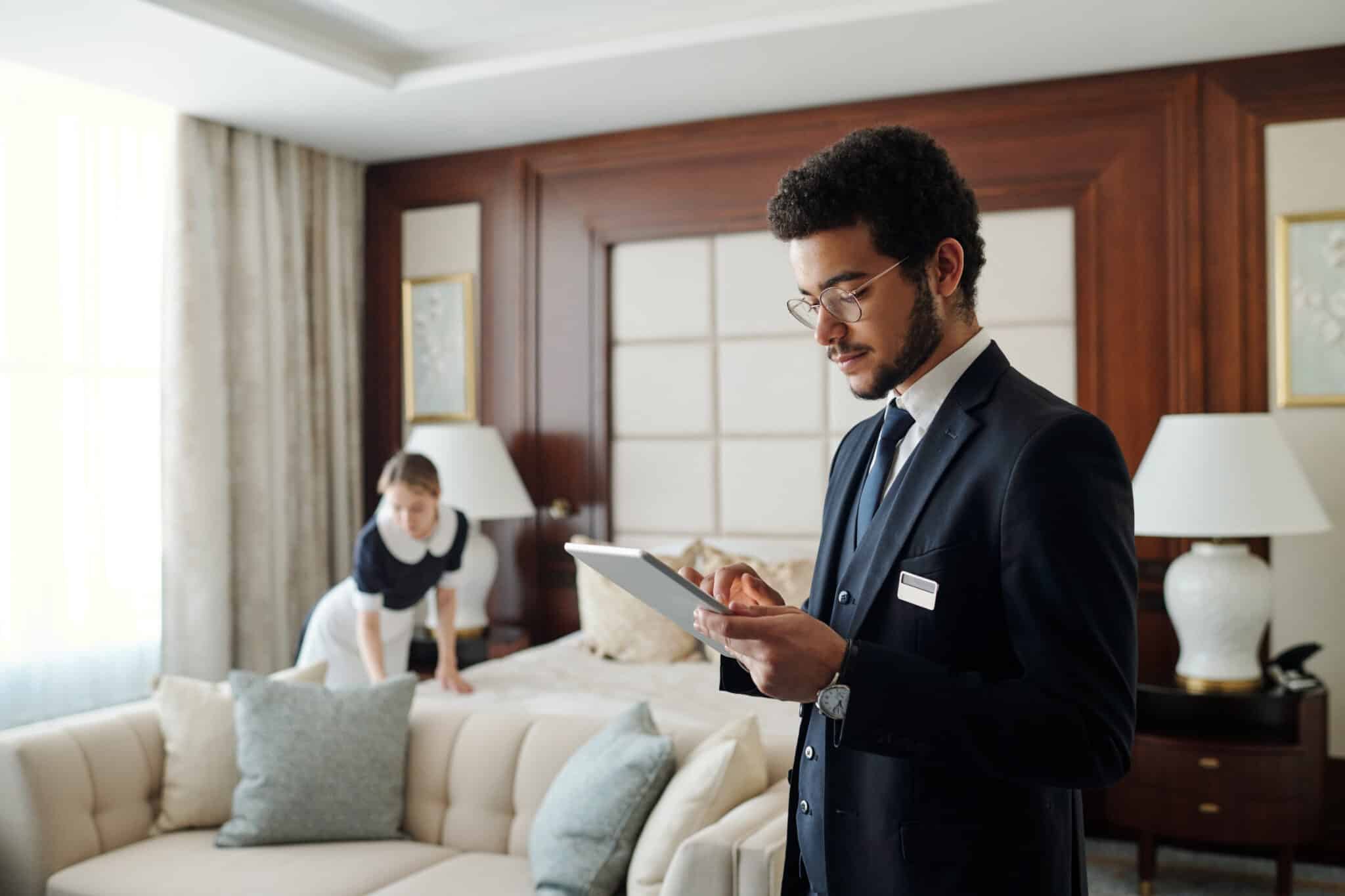 Young elegant entrepreneur in formalwear using tablet while standing in front of camera in hotel room with chambermaid on background
